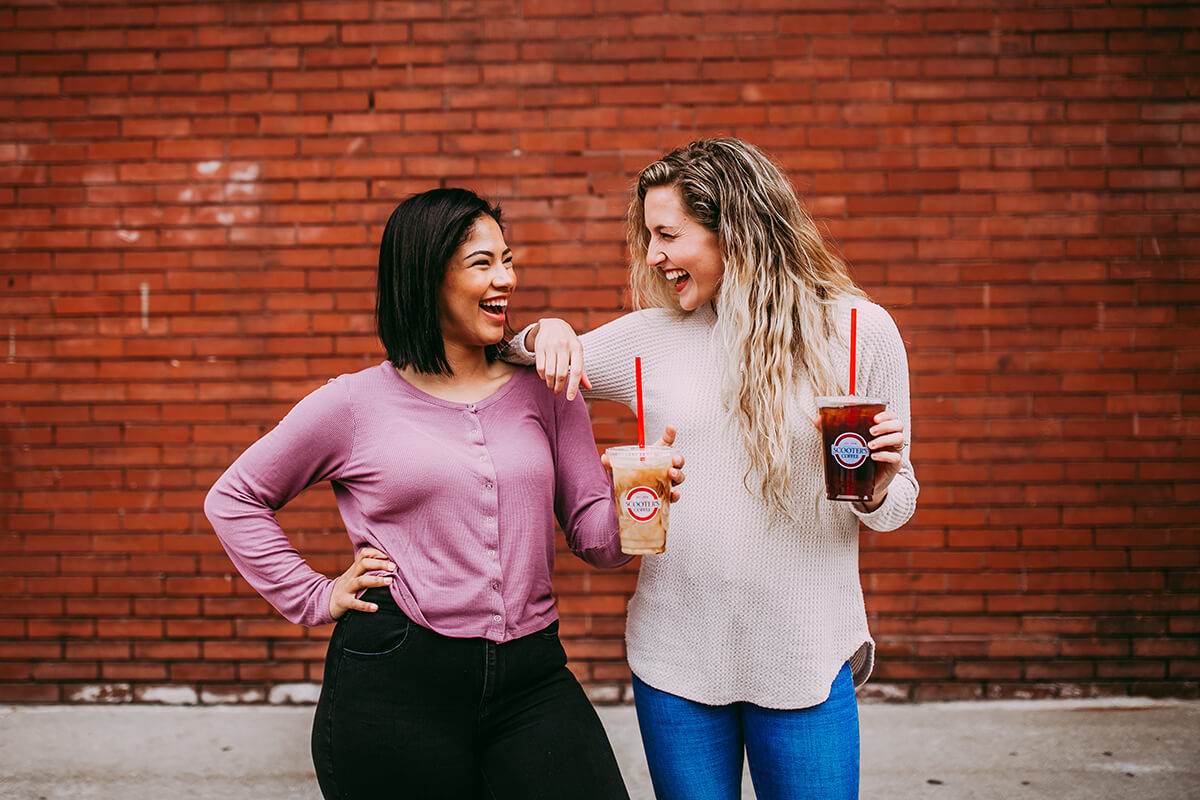 Two women laughing and holding coffee in front of brick wall