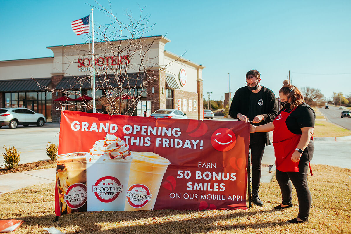 Two people standing near a sign that says Grand Opening This Friday Earn 50 Bonus Smiles on our Mobile App